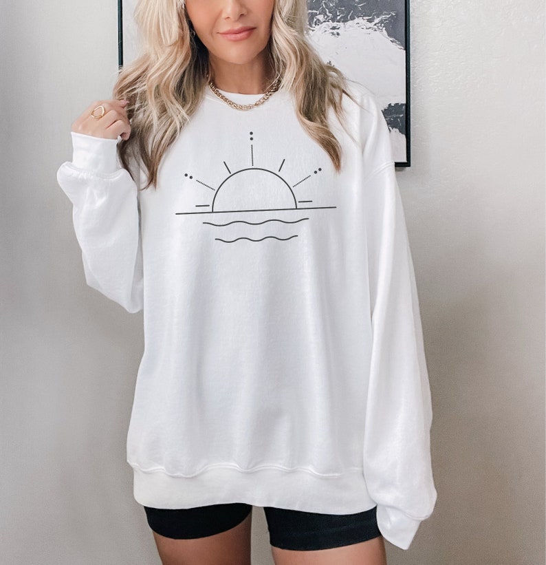 May include: White sweatshirt with a black line drawing of a sun setting over wavy water.