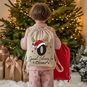 A child wearing a Santa hat and carrying a red bag stands in front of a Christmas tree, with gift boxes visible in the background.
