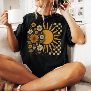 A woman sitting on a couch, holding a white mug and wearing a black t-shirt with a sun and flower design.
