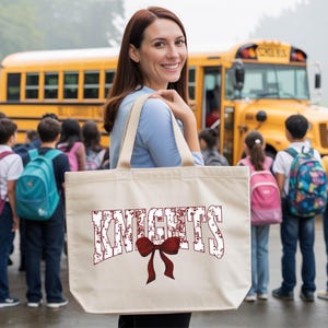 A woman holding a large tote bag with the word "KNIGHTS" printed on it, standing in front of a group of students and a yellow school bus.