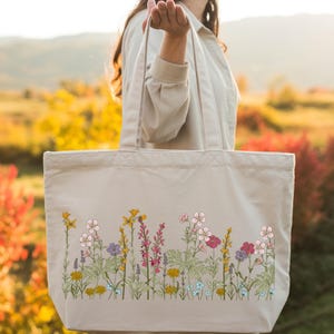 A woman is holding a large tote bag with a floral pattern on it, standing in a field with colorful flowers in the background.