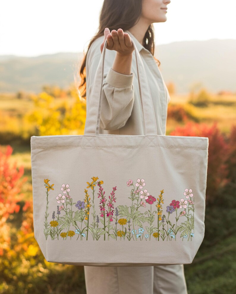 A woman is holding a large tote bag with a floral pattern on it, standing in a field with colorful flowers in the background.