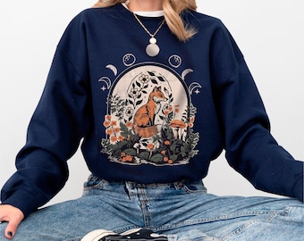 Puede incluir: Una sudadera azul marino de cuello redondo con un estampado gráfico central de un zorro naranja sentado entre flores, helechos y setas dentro de un marco arqueado, rodeado de fases lunares y estrellas, combinada con pantalones vaqueros azules.