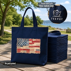 May include: A navy blue tote bag featuring a rectangular patch with a patchwork-style American flag design in red, white, and blue. A stack of identical navy blue tote bags sits beside it. An inset shows a white teapot logo and the text TEEPOT.