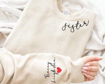 May include: A beige crewneck sweatshirt featuring the word sister in black cursive script on the upper chest. The sleeve displays the name Tracey Leigh Ann in black cursive script accompanied by a small red heart graphic near the wrist cuff.