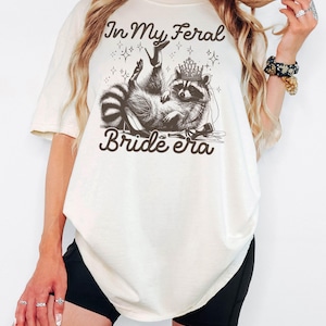 May include: Cream-colored t-shirt featuring a raccoon wearing a crown and veil, reclining with high heels. The text "In My Feral Bride Era" is printed above and below the raccoon illustration.