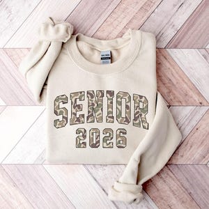 May include: A beige crewneck sweatshirt with the word "SENIOR" and the number "2026" printed on the front in a camouflage pattern. The sleeves are folded up. The sweatshirt is made of a soft material.