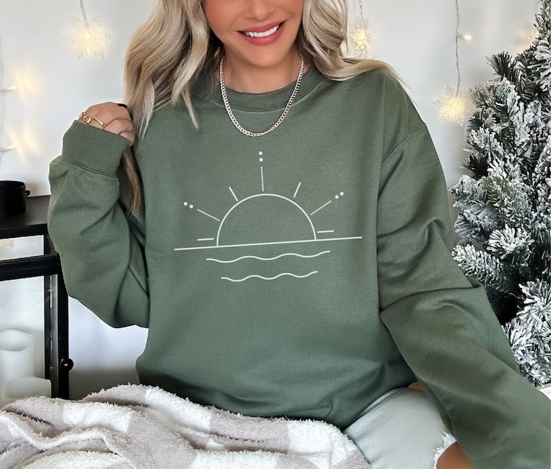 May include: A green sweatshirt featuring a white graphic of a sun setting over water. The graphic is a simple line drawing with a circle for the sun and wavy lines for the water.