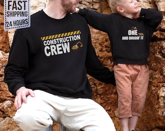 May include: Two black long-sleeve sweatshirts. The larger sweatshirt features yellow and black caution stripes with the text CONSTRUCTION CREW and an excavator graphic. The smaller sweatshirt features the text ONE AND DIGGIN' IT with an excavator.