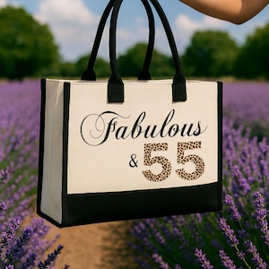 A hand holding a tote bag with the text "Fabulous & 55" printed on it, standing in front of a field of purple flowers.