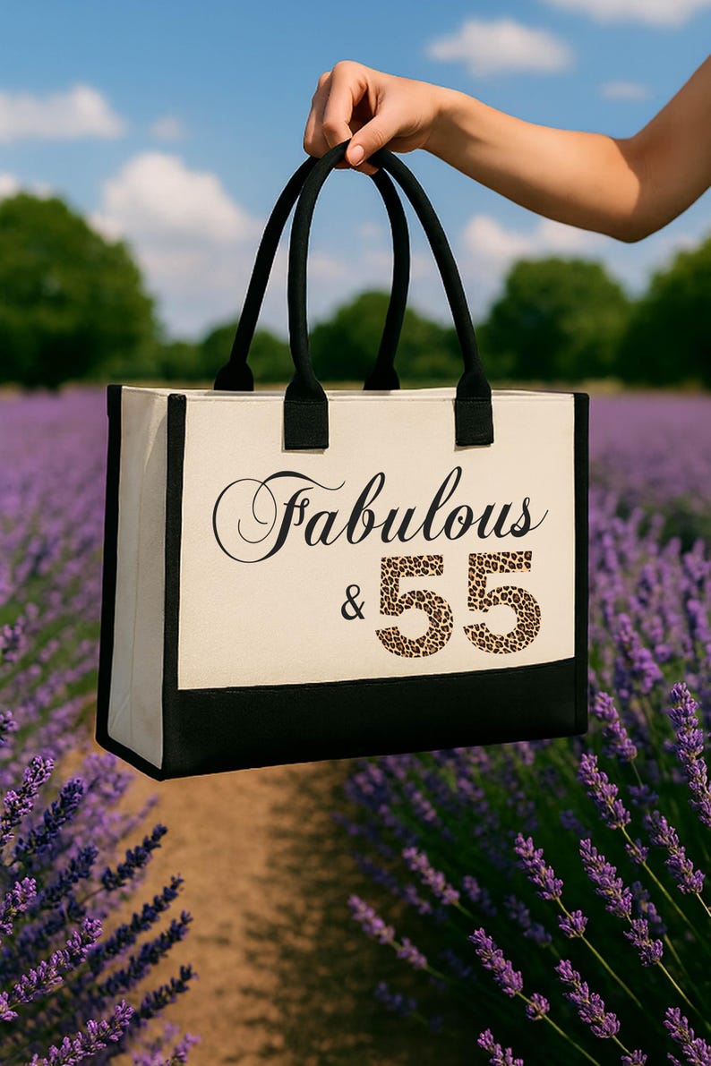 A hand holding a tote bag with the text "Fabulous & 55" printed on it, standing in front of a field of purple flowers.