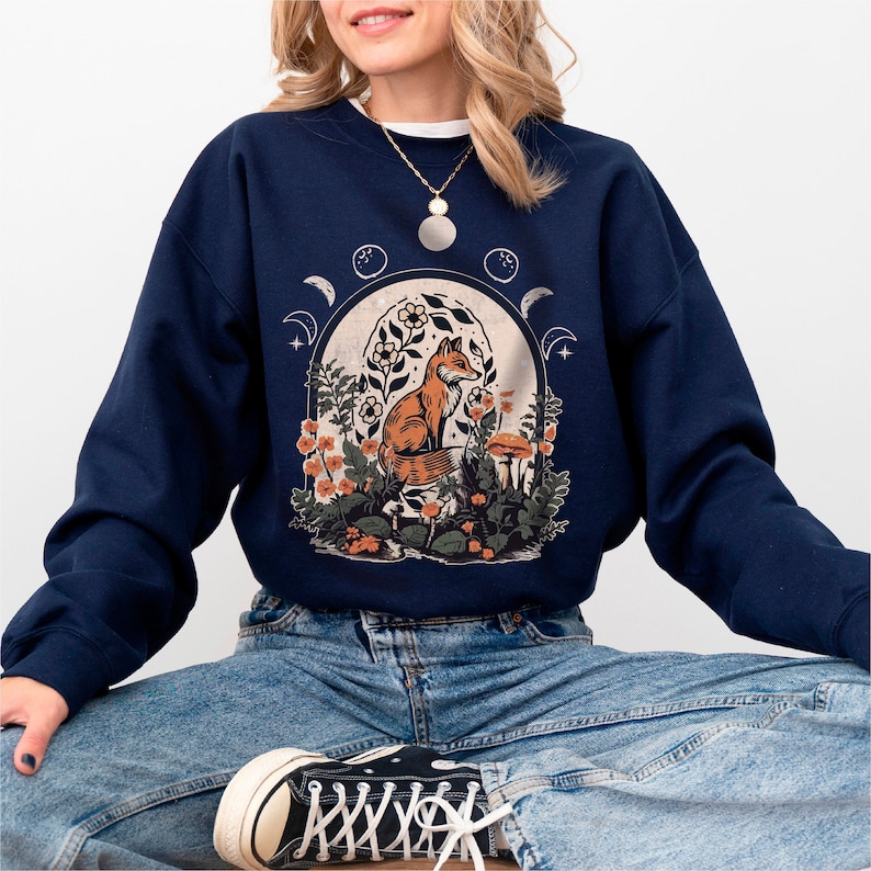 May include: A navy blue sweatshirt with a graphic of a fox sitting in a moonlit forest with flowers and mushrooms. The sweatshirt has a crew neck and long sleeves.