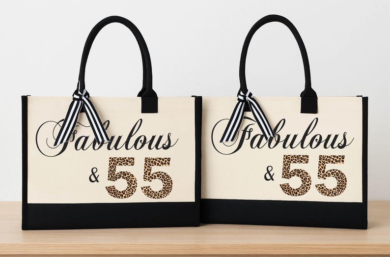Two beige tote bags with black handles and ribbons, each with the text "Fabulous & 55" printed on them.