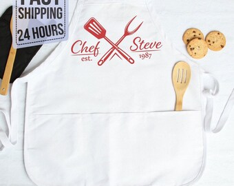 May include: A white apron featuring a red graphic of a crossed spatula and fork with the text Chef Steve est. 1987. The apron includes a front pocket, a wooden spoon, and two round chocolate chip cookies placed on the fabric surface.