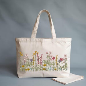 A white tote bag with a floral embroidered design on the front, featuring various colorful flowers and plants.