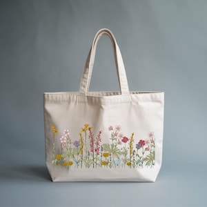 A white tote bag with a floral embroidered design on the front, featuring various colorful flowers and plants.