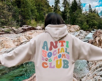 May include: A beige hooded sweatshirt featuring a multi-colored graphic on the back with the text ANTI SOCIAL DOG MOMS CLUB in a wavy, retro-inspired font. The design includes a small paw print icon integrated into the colorful lettering.