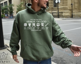 Op de afbeelding: Een olijfgroene hoodie met witte tekst "PUT ON THE FULL ARMOR OF GOD" boven iconen die de gordel van waarheid, het borstpantser van gerechtigheid, de schoenen van vrede, het schild van geloof, de helm van redding en het zwaard van de geest voorstellen.