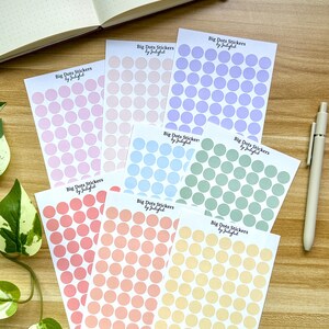 May include: A collection of dot sticker sheets in various pastel colours, including pink, peach, blue, green, orange, and yellow. Each sheet is labelled "Big Dots Stickers by Jubiful". A pen and notebook are also present.