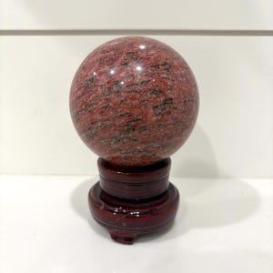 May include: A red granite sphere with black veining, displayed on a dark red wooden stand. The polished stone ball has a smooth surface and is a decorative item. The stand has a round base and a pedestal.
