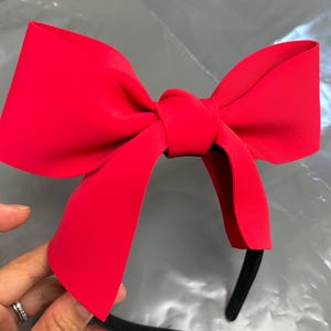 May include: A bright red bow headband. The bow is large and prominently displayed, with a smooth, matte finish. The headband itself is black and slender, providing a contrasting base for the vibrant bow. The accessory is held by a hand.