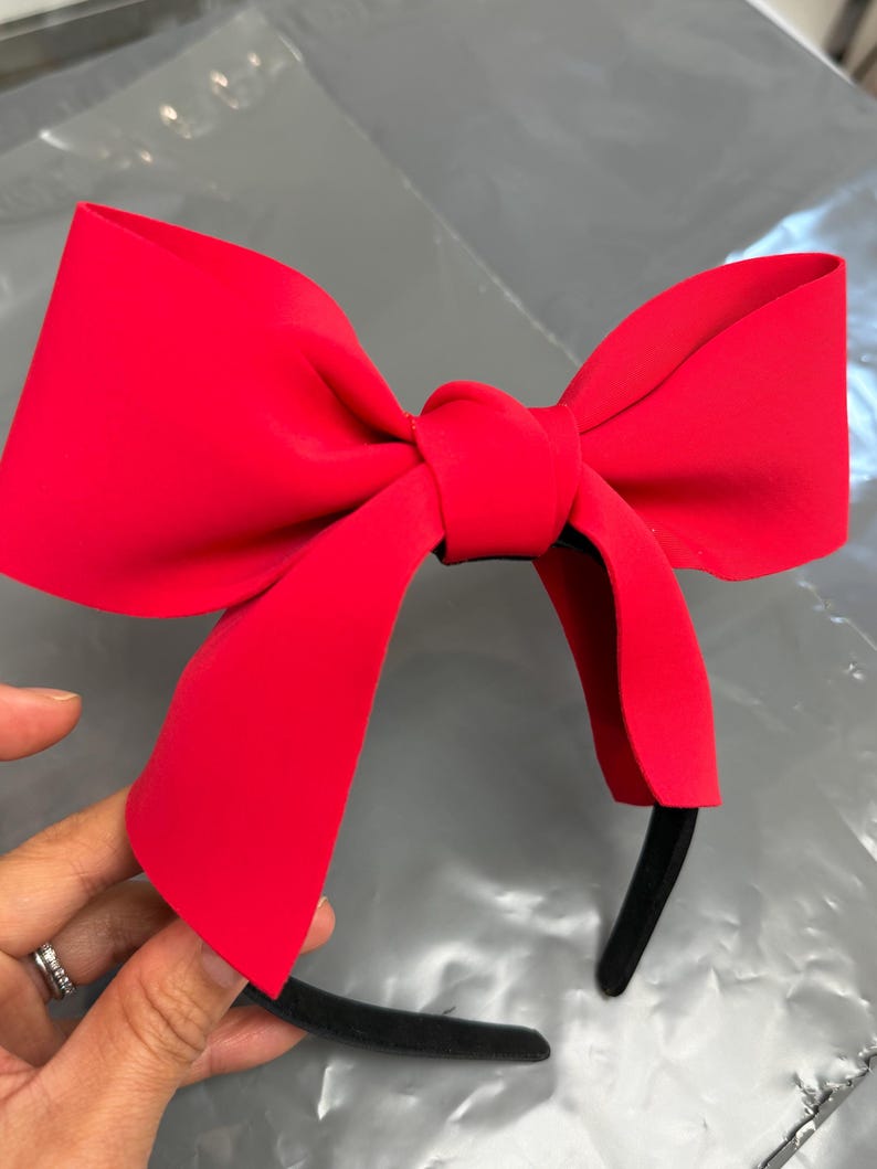 May include: A bright red bow headband. The bow is large and prominently displayed, with a smooth, matte finish. The headband itself is black and slender, providing a contrasting base for the vibrant bow. The accessory is held by a hand.