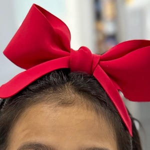 May include: A vibrant red bow headband. The large bow is centered atop the head, with the bow's fabric appearing smooth and slightly reflective. The headband is a solid color, perfect for adding a pop of color to any outfit.