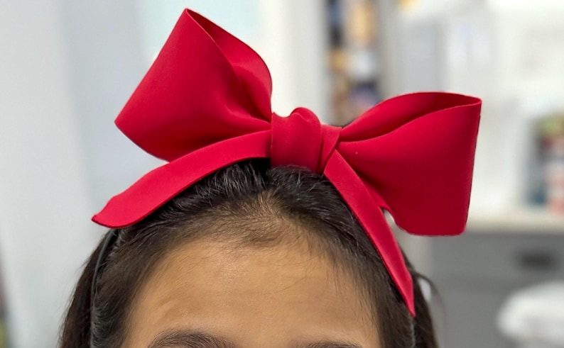 May include: A vibrant red bow headband. The large bow is centered atop the head, with the bow's fabric appearing smooth and slightly reflective. The headband is a solid color, perfect for adding a pop of color to any outfit.