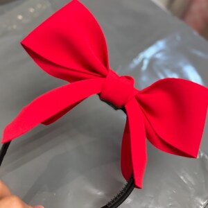 May include: A bright red bow headband. The bow is large and made of a smooth fabric. The headband is black and has a simple design. The bow is centered on the headband.