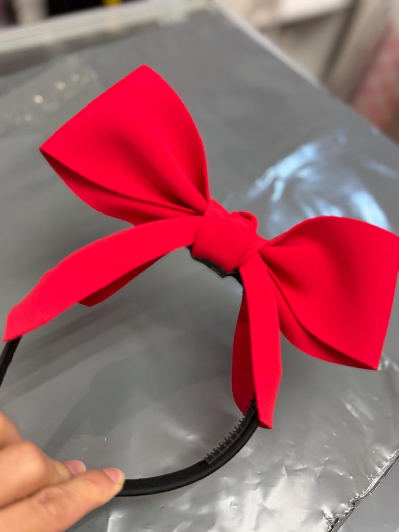 May include: A bright red bow headband. The bow is large and made of a smooth fabric. The headband is black and has a simple design. The bow is centered on the headband.