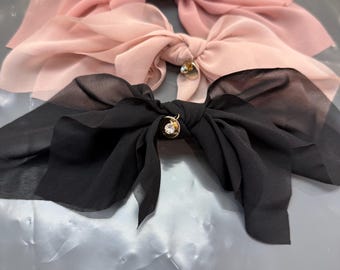 May include: Three sheer fabric hair bows in shades of pink and black. Each bow features a delicate, flowing design and is accented with a small, gold-colored embellishment. The bows are arranged on a reflective surface.