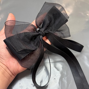 Pode incluir: Um laço de organza preto com fitas longas e esvoaçantes. O laço é amarrado no centro e possui várias camadas de tecido transparente, criando um efeito volumoso. As fitas são feitas de um material semelhante e são longas o suficiente para amarrar ou drapejar.