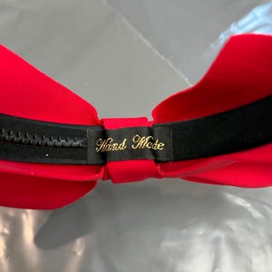 May include: A red strapless bra with a black band and zipper closure. The interior is red, and a small black tag with gold text that reads "Hand Made" is visible.