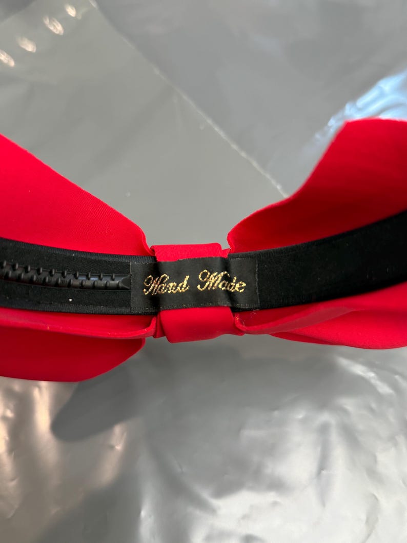 May include: A red strapless bra with a black band and zipper closure. The interior is red, and a small black tag with gold text that reads "Hand Made" is visible.