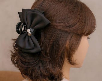 May include: A black bow hair clip with a pearl and rhinestone embellishment. The clip is attached to brown hair, and the bow is made of a soft, flowing fabric. The hair accessory is a stylish addition to any hairstyle.