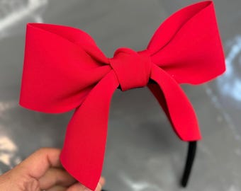 May include: A bright red bow headband. The large bow is the focal point, with a smooth, solid texture. The headband itself is black and thin, providing a contrasting base for the vibrant bow. This accessory is perfect for adding a pop of color.