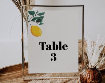 Puede incluir: Tarjeta de número de mesa enmarcada, con una ilustración de limón y el texto "Table 3". El marco dorado se encuentra sobre una bandeja de madera, complementado con elementos decorativos, creando una estética elegante.