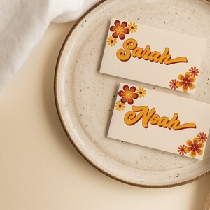 May include: Two place cards with the names "Sarah" and "Noah" written in a retro font. The cards are decorated with orange and yellow flowers.