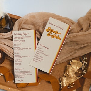 May include: Wedding ceremony programs with the names "Sarah & John" in retro font. The programs are cream-colored with orange and yellow accents. The programs are on a wooden tray with a candle, fabric, and decorative elements.