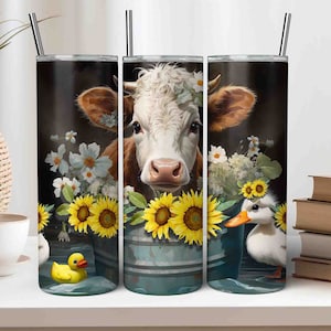 May include: Three stainless steel tumblers with a cow, sunflowers, and ducks design. The cow is white with brown spots and has a flower crown. The sunflowers are yellow with black centers. The ducks are white with orange beaks. The tumblers are on a white surface.