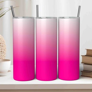 Könnte beinhalten: Drei Edelstahlbecher mit rosafarbenen Ombre-Designs und durchsichtigen Deckeln. Die Becher stehen auf einer weißen Oberfläche mit einer Pflanze und Büchern im Hintergrund.