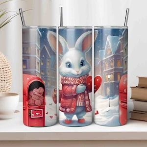 Op de afbeelding: Drie roestvrijstalen bekers met een winterscène met een wit konijn dat een rood hart vasthoudt. Het konijn draagt een rood-witte trui. De scène bevat ook een rode brievenbus met harten erin en een besneeuwde straat.