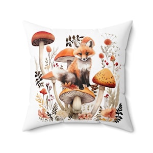 May include: A white throw pillow with a watercolor illustration of a fox sitting on a cluster of mushrooms. The mushrooms are orange and brown with white spots. The fox is orange and white with a bushy tail. The background is white with green leaves and flowers.