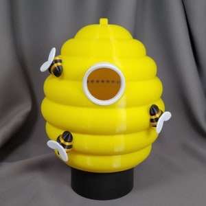 Puede incluir: Un recipiente de cerámica amarillo con forma de colmena con una abertura redonda y tres abejas negras y amarillas adheridas. El recipiente tiene una base negra.