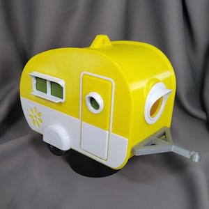 Puede incluir: Una caravana de juguete amarilla y blanca con un detalle de margarita, una ventana pequeña y una ventana redonda con un toldo blanco. La caravana tiene un enganche de remolque y una rueda negra. Un artículo decorativo.