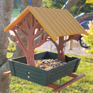 Puede incluir: Un comedero para pájaros con un techo de madera marrón y una base verde. El comedero está lleno de semillas para pájaros y tiene un diseño de árbol decorativo.
