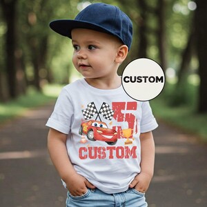 Puede incluir: Un ni&ntilde;o peque&ntilde;o con una gorra azul marino y una camiseta blanca con un gr&aacute;fico de coche de carreras rojo. La camiseta lleva la palabra "CUSTOM" y banderas a cuadros. El ni&ntilde;o est&aacute; de pie en un camino pavimentado.