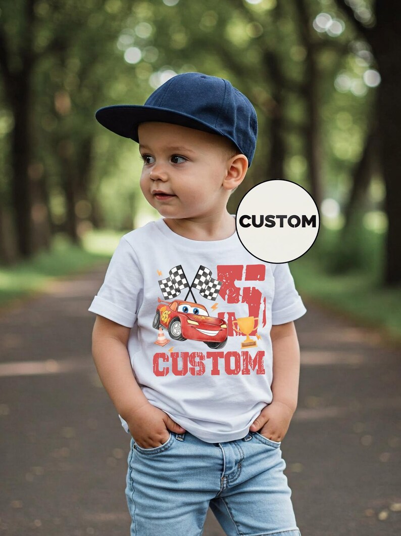 Puede incluir: Un ni&ntilde;o peque&ntilde;o con una gorra azul marino y una camiseta blanca con un gr&aacute;fico de coche de carreras rojo. La camiseta lleva la palabra "CUSTOM" y banderas a cuadros. El ni&ntilde;o est&aacute; de pie en un camino pavimentado.
