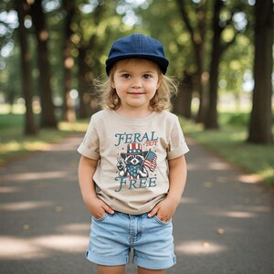 May include: A child wearing a tan t-shirt with the words "FERAL BUT FREE" and a raccoon graphic in a patriotic design. The child is also wearing a navy blue cap and denim shorts.