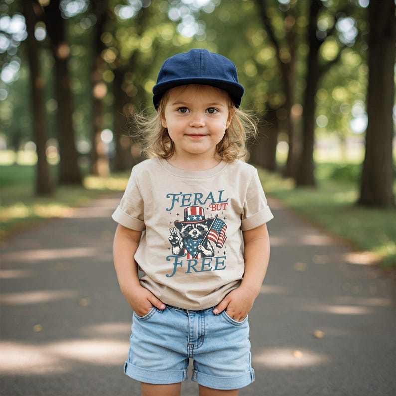 May include: A child wearing a tan t-shirt with the words "FERAL BUT FREE" and a raccoon graphic in a patriotic design. The child is also wearing a navy blue cap and denim shorts.